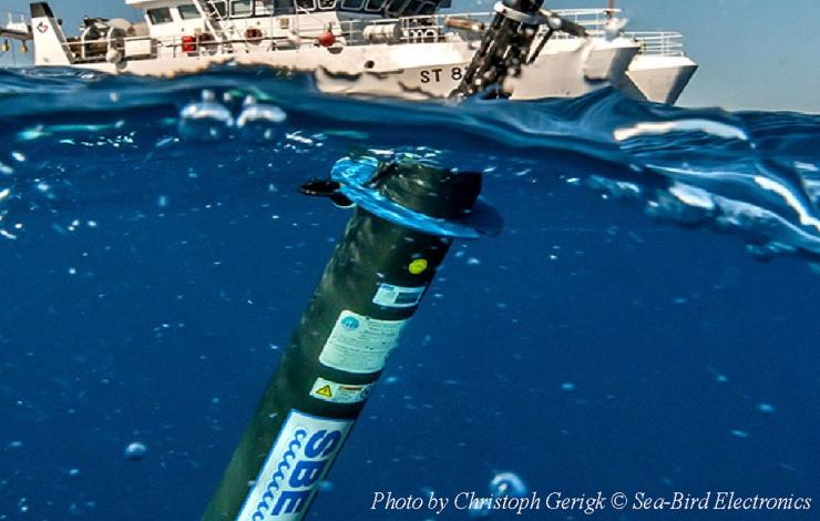 Underwater image of a long cylindrical tube with a sensor on top floating above the water with a ship in the background