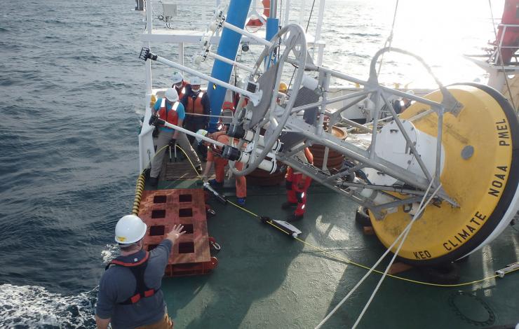 Folks on the back deck of a ship in a half circle around a large yellow buoy with metal cage signaling to each other in preparation to deploy the buoy off the back of the ship