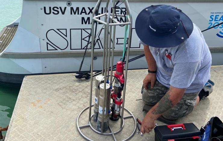 A person with a blue rimmed hat with gray shirt and camouflage pattern shorts kneeling in front of a metal cage hosting 3 silver instruments with a toolbox nearby in front of the uncrewed surface vessel with water behind. 