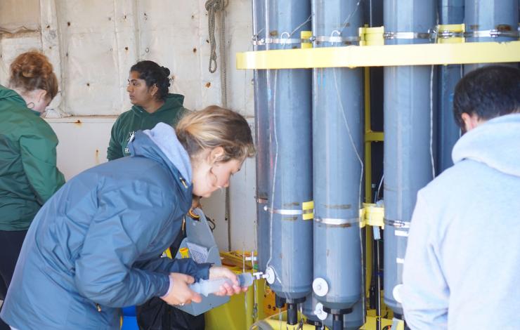 People bent over slightly around a CTD rosette of grey niskin bottles collecting water samples using a syringe through a valve