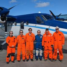 The aerial flight team of six pose in front of a blue and white NOAA Twin Otter aircraft wearing flight suits in both orange and blue. 