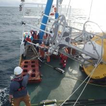 Folks on the back deck of a ship in a half circle around a large yellow buoy with metal cage signaling to each other in preparation to deploy the buoy off the back of the ship