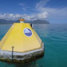 Yellow octagonal shaped buoy in greenish-blue clear water with clouds i the sky and land in the background