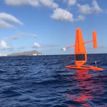 Orange drone with sail in open water in front of some bluffs and coastal city  in the distance