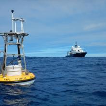 Yellow buoy floating on top of the water with a metal cage above with various instruments with a ship in the background