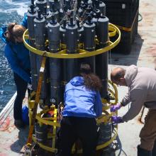 People stand around a circular CTD instrument collecting water samples on a ship out in the Pacific Ocean