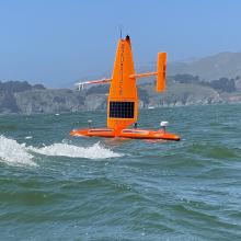 orange sailing drone in on the water with some waves sailing in front of San Francisco bluffs