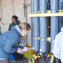 People bent over slightly around a CTD rosette of grey niskin bottles collecting water samples using a syringe through a valve